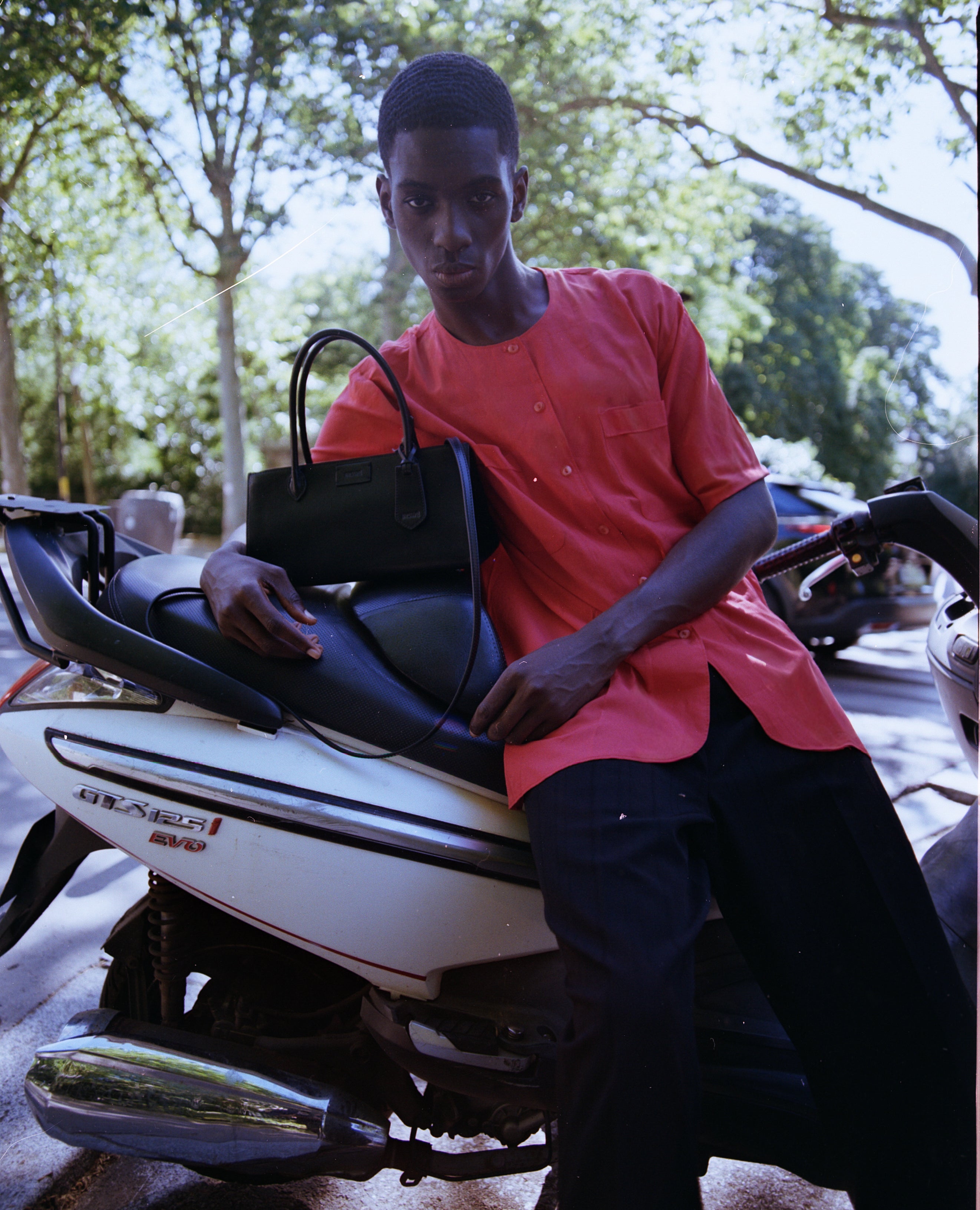 A man wearing a coral-red short-sleeve button-up shirt and black trousers sits on a white scooter outdoors, holding a black INSWE CESARINE GRANDE leather handbag. Trees and a street are visible in the background.